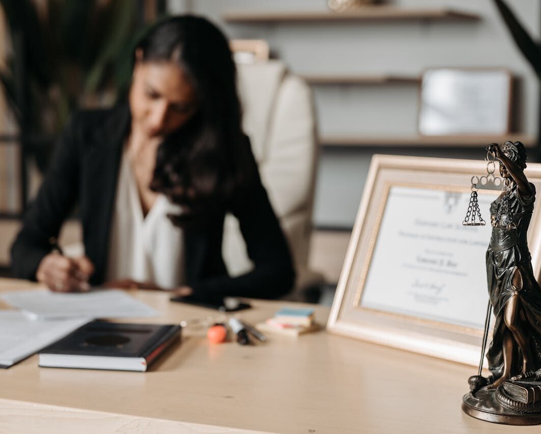 Focused woman writing at desk with Lady Justice figurine and certificate in office.